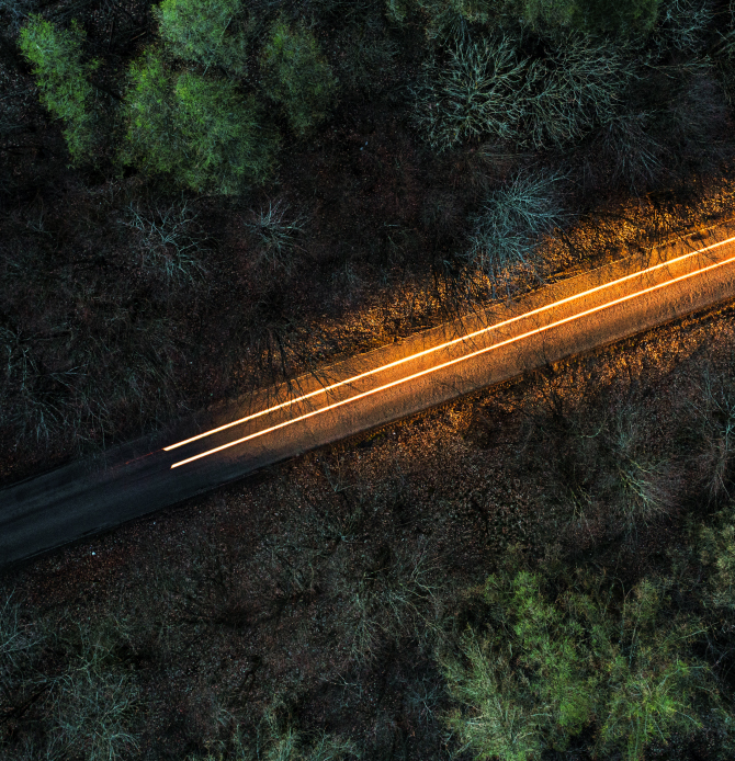 Aerial view of a road with a vehicle's light trail through a dark forest.
