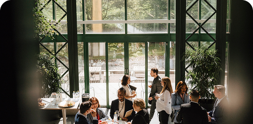 A group of people in business attire converse in a well-lit room with large windows and visible greenery outside.